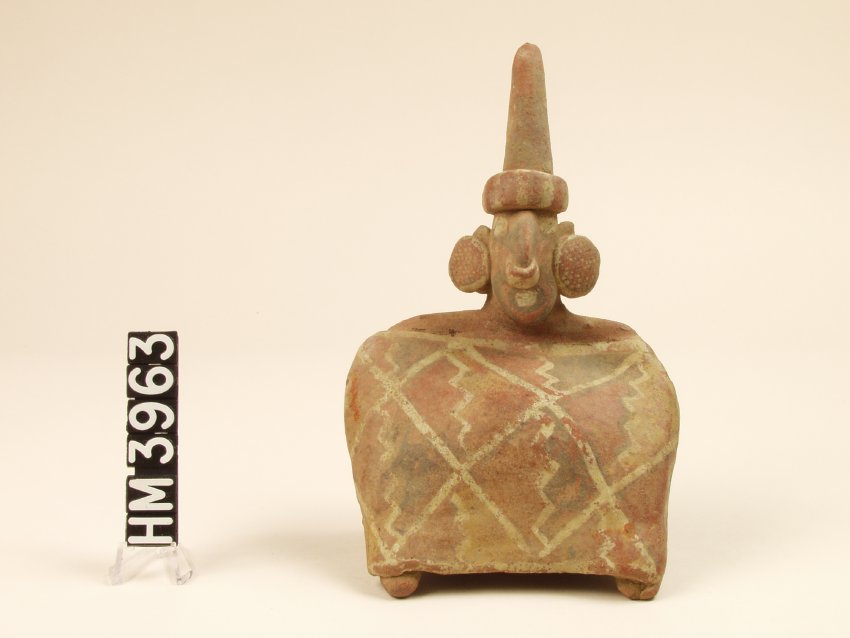 Small ceramic figure. Body is a squat rectangle with a head at center top. Ear spools and large, pointed headdress.