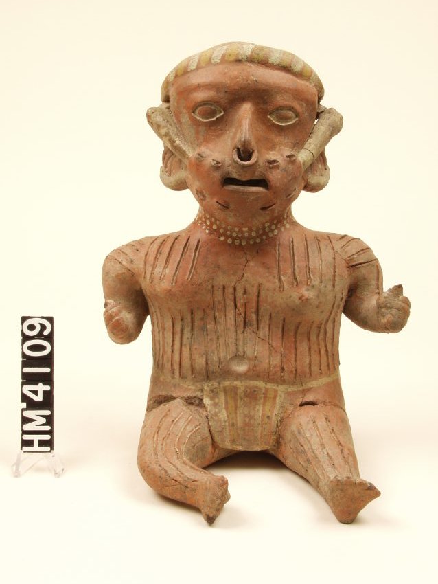 Ceramic figure with large head with a white and yellow striped headband. Carved oval eyes filled with white paint, a pointed nose with a large nose ring, and small ears with large white earrings. Mouth is open with a white object sticking out of each cheek, with stitches visible. White painted dot necklace and body is scored vertically. Figure is wearing a waist garment. Fists are clenched with bent elbows to side and legs are in front of body.