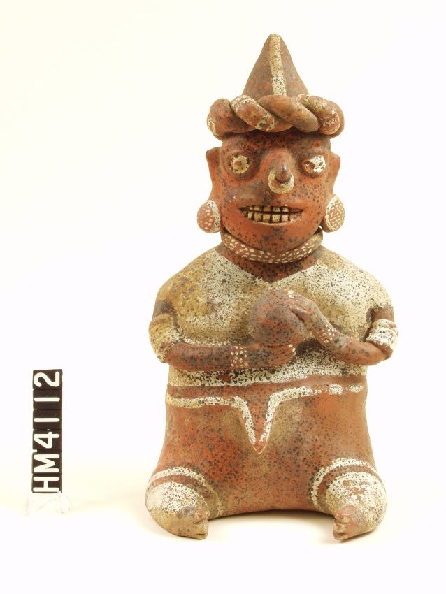 Seated ceramic figure with short legs. Thin arms bent 90 degrees at elbow holding ball, left hand on top of ball, right cradling it. Eyes almond-shaped, painted white. Large nose with nose ring painted white. Disc earrings with white rim and dots, necklace with white dots. White short sleeved garment. Three to five white bands on each arm, four strand beaded bracelet on each wrist. Fingernails painted white. Narrow white band on upper legs and broad white band covering most of legs, painted. Wearing pointy hat with double twisted horizontal band, one plain red, one painted with white stripes. Vertical white stripe down middle of hat.