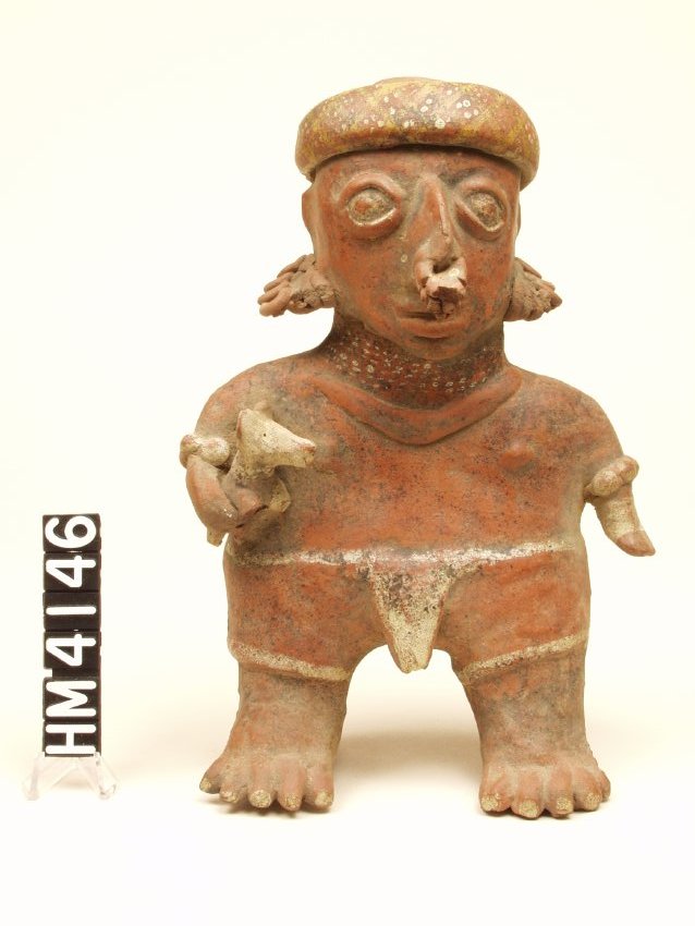 Standing ceramic figure with stylized short arms, left hanging at side and right holding a dog. Modeled almond eyes in white, modeled lips, large nose with three incised white modeled rings. Broad flat headband with yellow cross-hatching and white dots grouped in the diamonds. Six strand white bead necklace over one modeled necklace. Disc arm band on each arm. Lower left arm painted white.