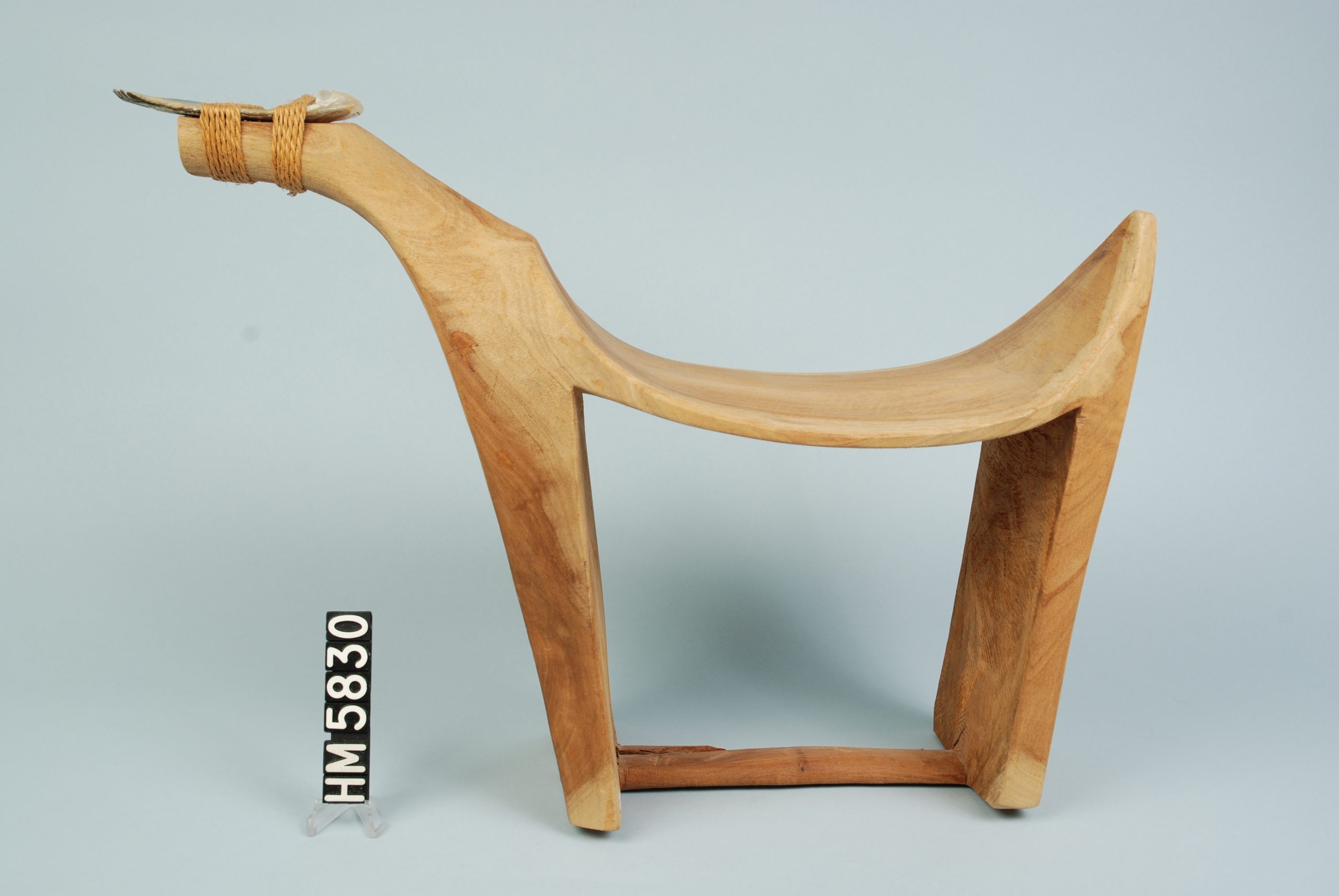 Image of an object formed as a seat that can be used with a leg on each side. Object is seen from the side. The front of the chair, seen to the left, sports a projection to which a sharpened blade is lashed.