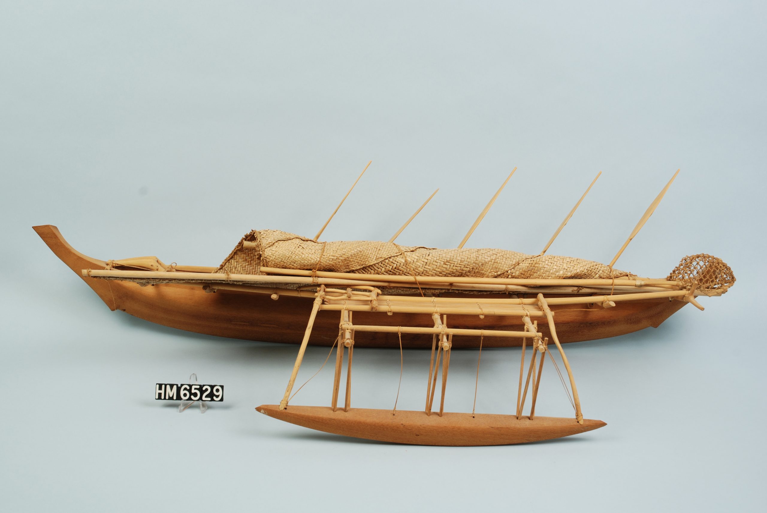 Image of boat model. Boat is in the form of a long, wide canoe with outrigger and a sail furled on deck.