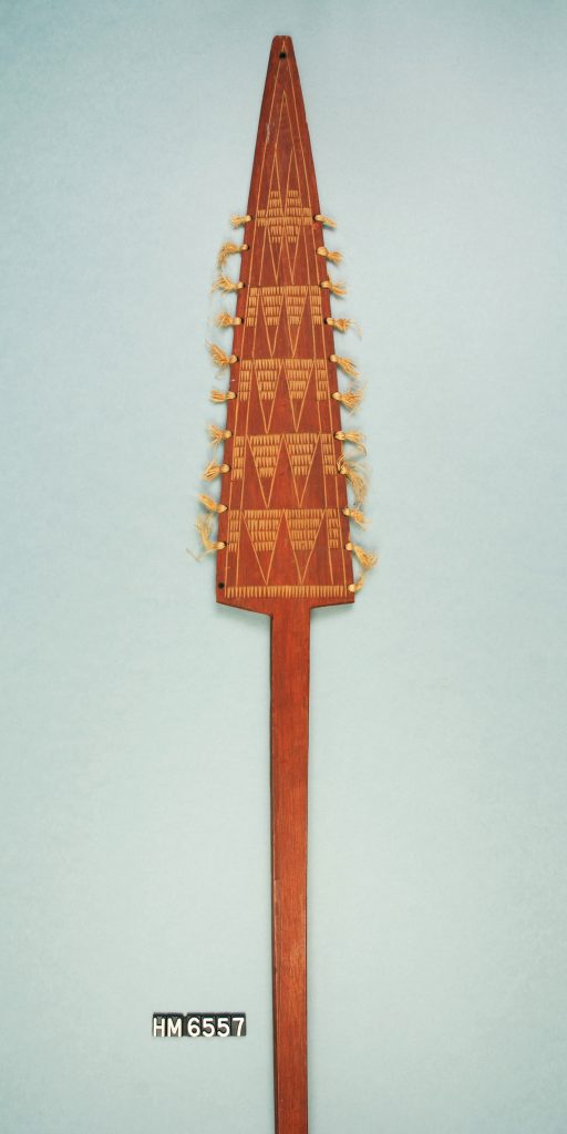 Image of a decorative paddle with head shaped like elongated triangle. Head is carved with triangular patterns and decorated with tufts of fiber.