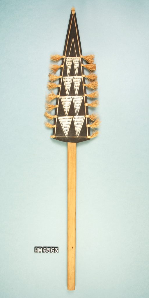 Image of a decorative paddle with head shaped like elongated triangle. Head is painted with black and white triangular patterns and decorated with tufts of fiber.