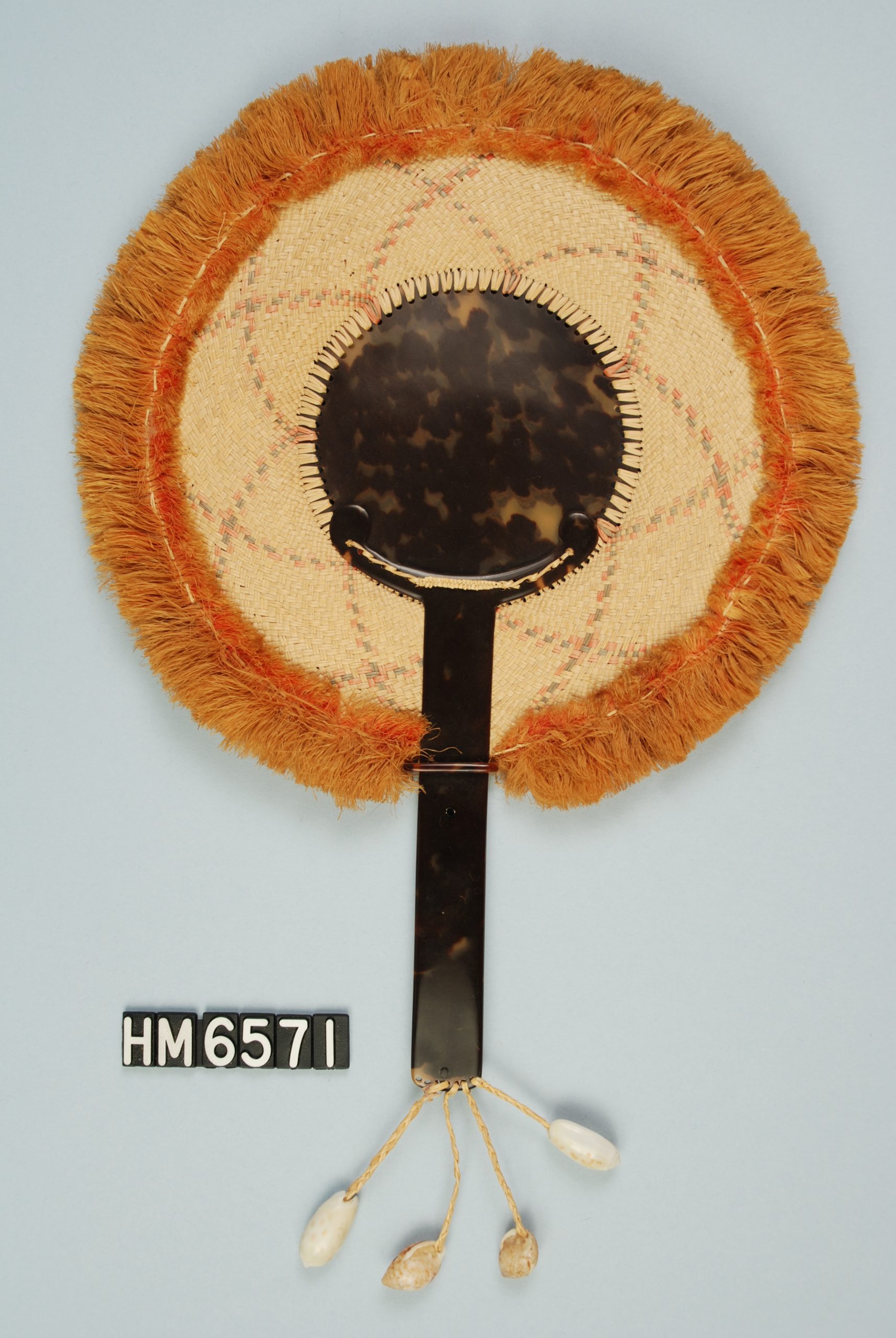 Image of a hand-held fan with circular head. Handle and center of head are tortoiseshell. Surrounding the center is an intricately woven strip attached to a fiber fringe at the edge.