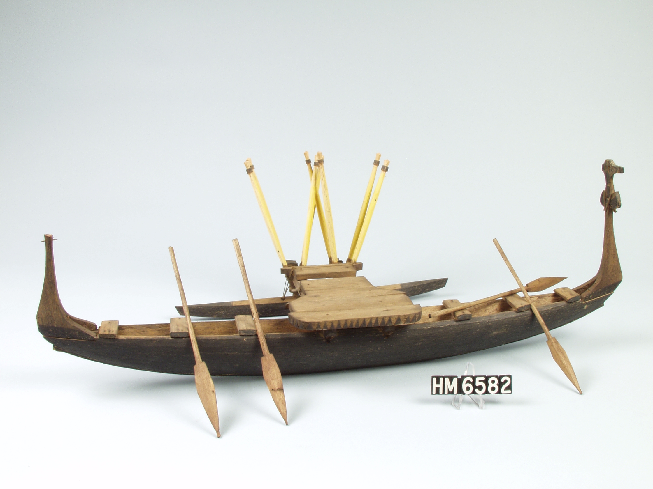 Image of a model boat, shaped long and narrow like a canoe with ends that come up into upright points. A platform is across the middle. Has tiny paddles.