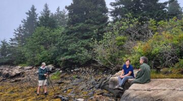 A photo of two people being recorded on video while sitting on a rock on the Maine coast