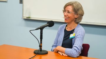 A photo of a woman sitting at a table next to a microphone.