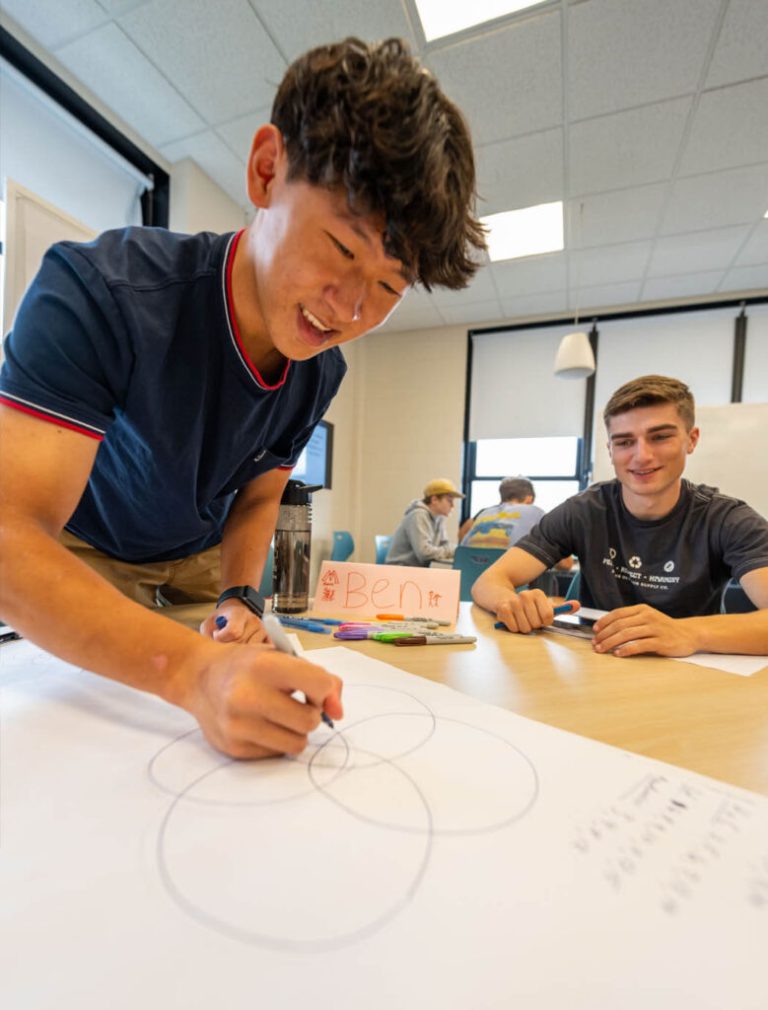 Image of students in a classroom. A young man is drawing a diagram on a piece of paper in the foreground.