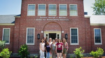 Charlotte Rhodes, MacKenzie Conant, Morelys Rodriguez Alfonso, Brooke MacDonald, Sara McBride, Shayla Miller, Molly Bennett and Jessica Beneski stand in front of Mitchell Center.