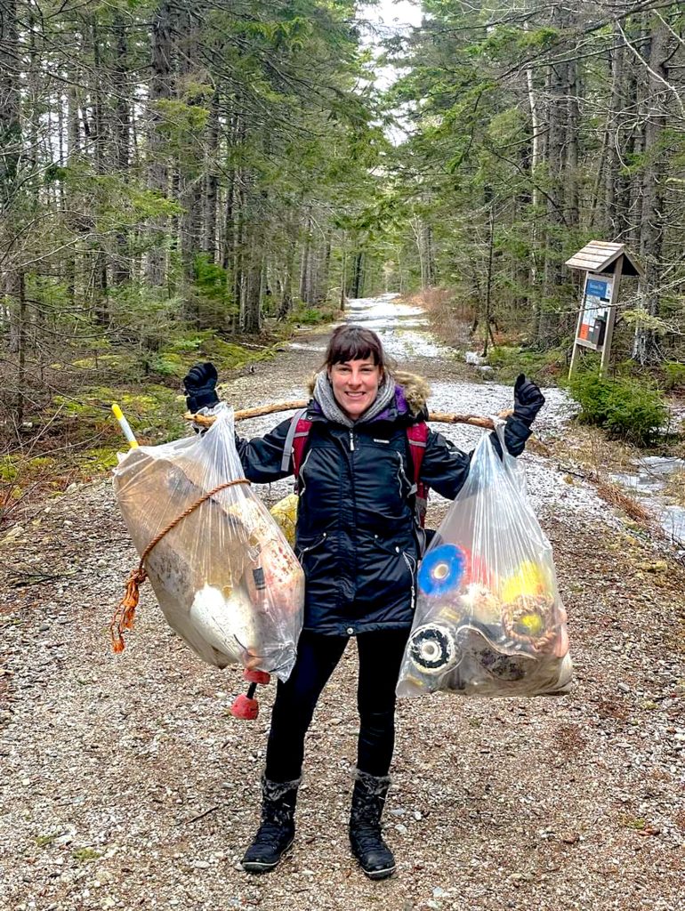 Cavalcanti stands in woods with stick over shoulders with a trash bag hanging from each end with material for sculpture