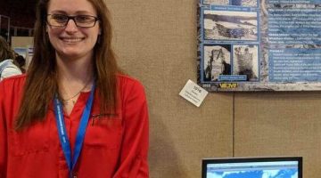Image of Emily Blackwood standing in front of her Presentation at the UMaine Student Symposium. She is wearing a red long sleeve shirt.